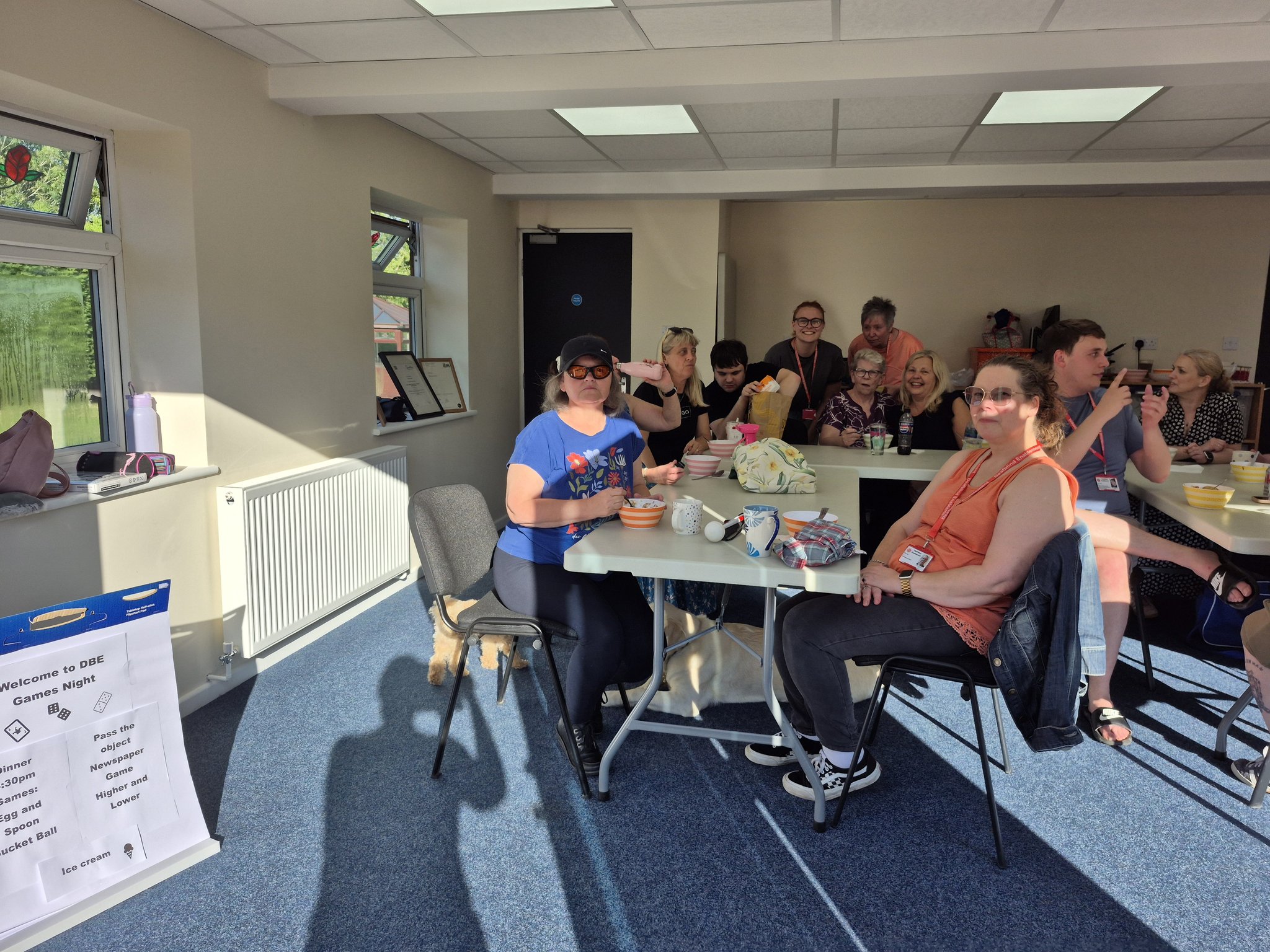 Service users and Communicator-Guides sitting around tables at Deafblind Enablement's Head Office, eating and chatting. A poster on the wall reads "Welcome to DBE Games Night" and lists the evening's games.