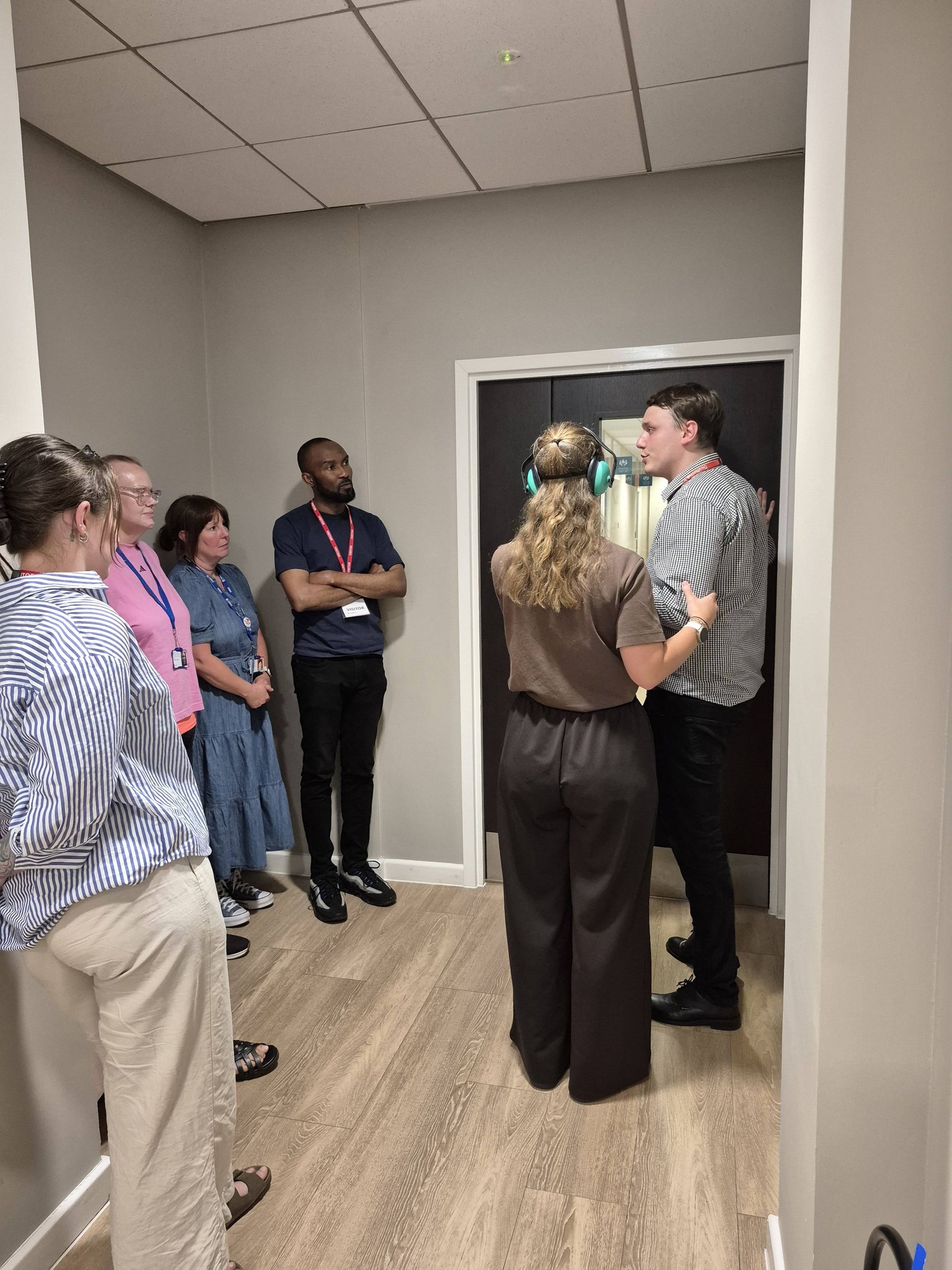 Students on a Signature Level 2 course watching a guiding demonstration in a corridor. One student wears ear defenders to simulate hearing loss while a trainer guides her through a doorway.